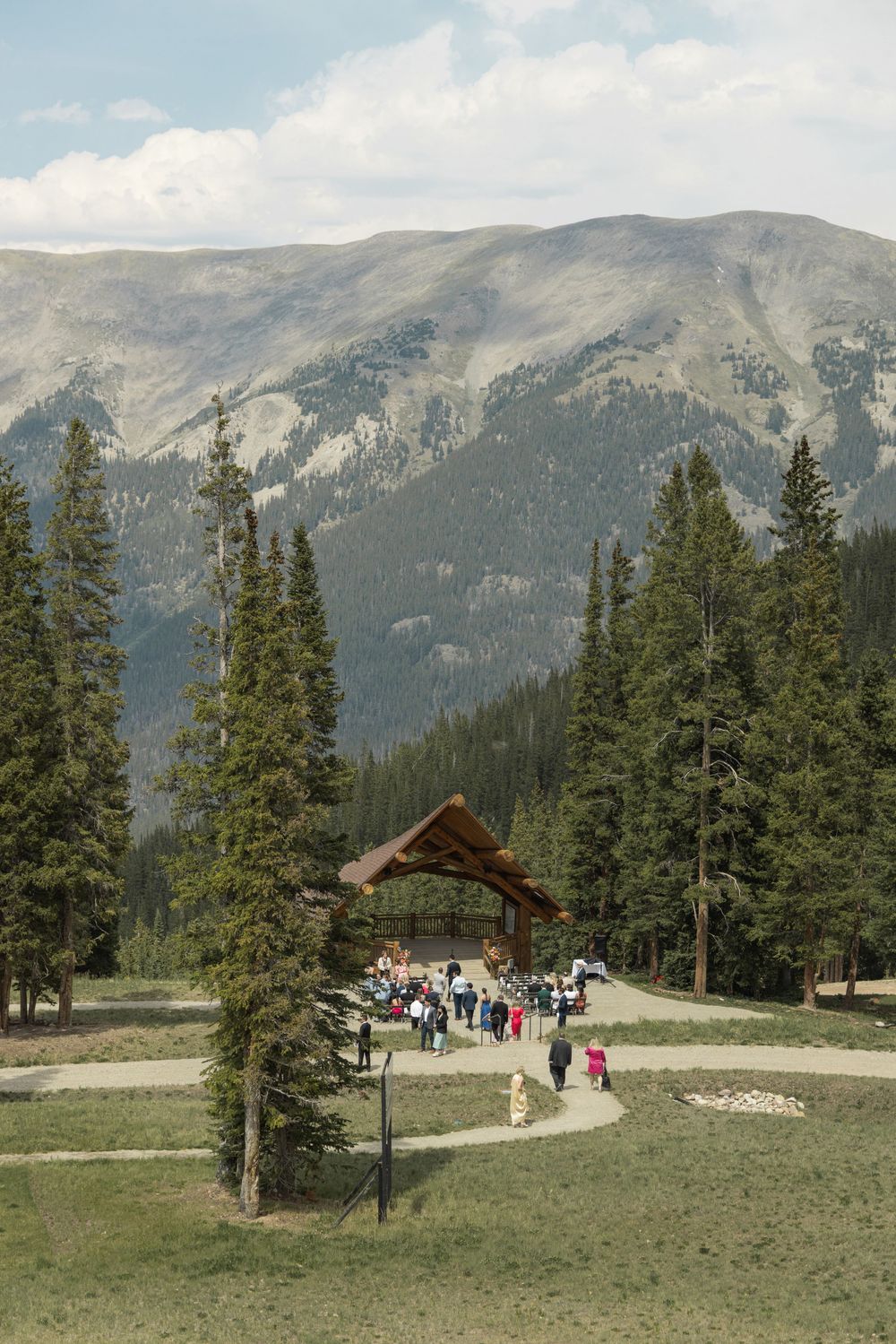 Outdoor mountain wedding ceremony with guests gathered under a wooden pavilion surrounded by pine trees and scenic peaks.
