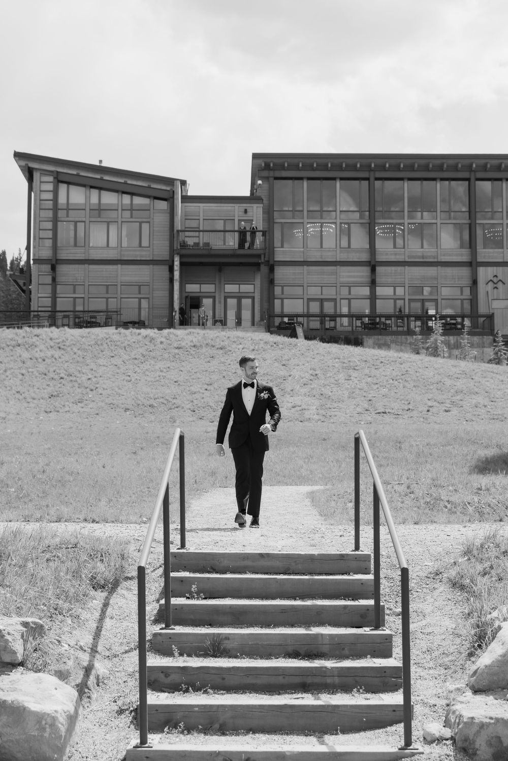 Groom in tuxedo descending outdoor stairs with large building backdrop in black and white wedding photo.