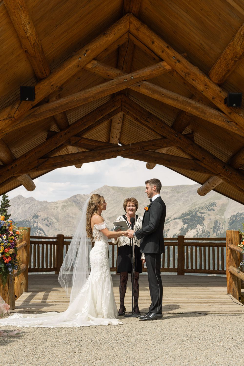 Bride and groom exchange vows under rustic wooden pavilion with mountain views during outdoor wedding ceremony.
