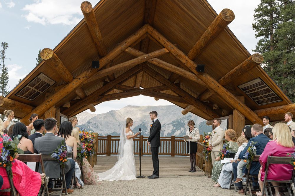 Outdoor mountain wedding ceremony under rustic wooden pavilion with couple at altar and guests seated amid stunning rocky landscape.