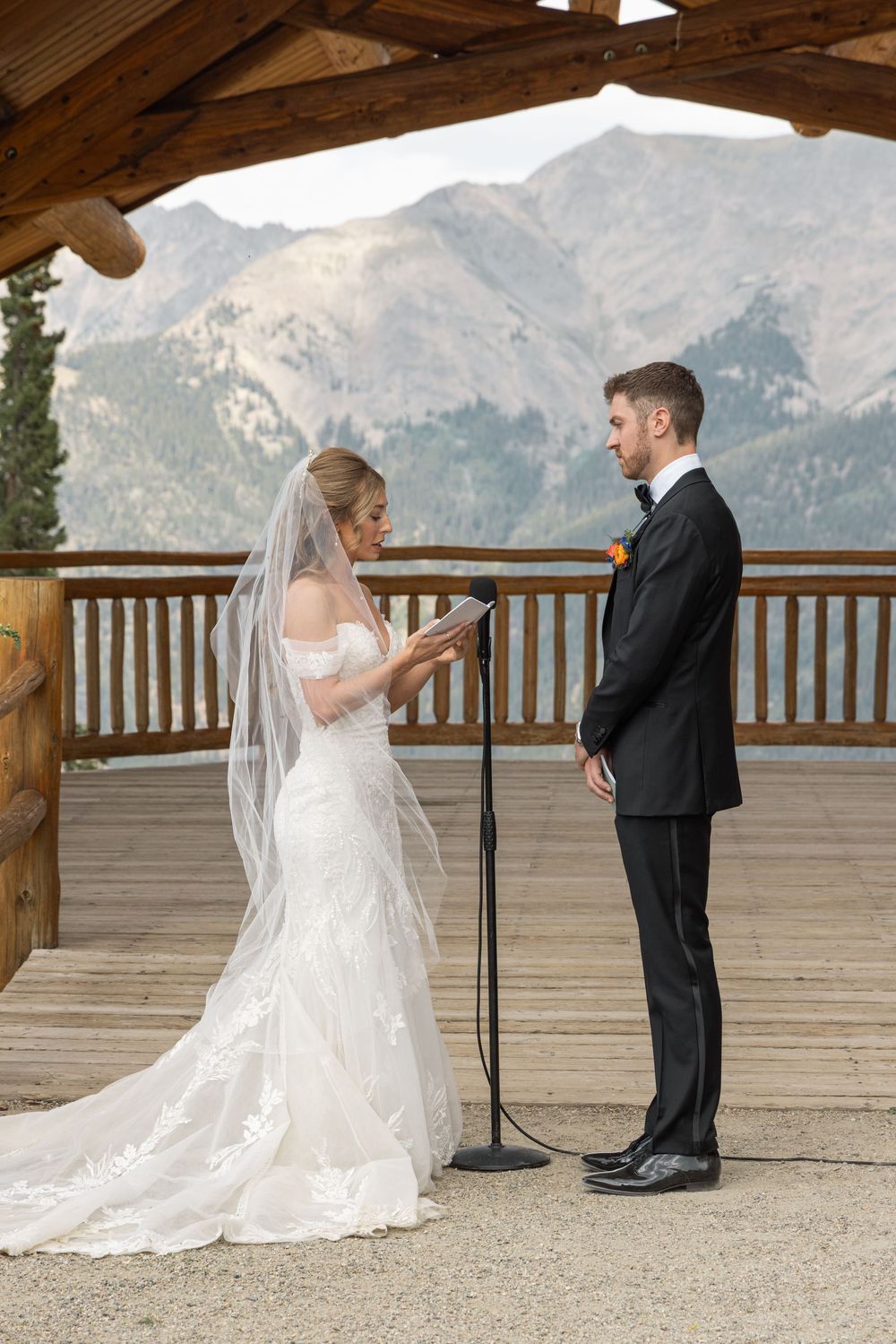 Bride reads vows to groom during outdoor mountain wedding ceremony on wooden deck with scenic rocky peaks backdrop.