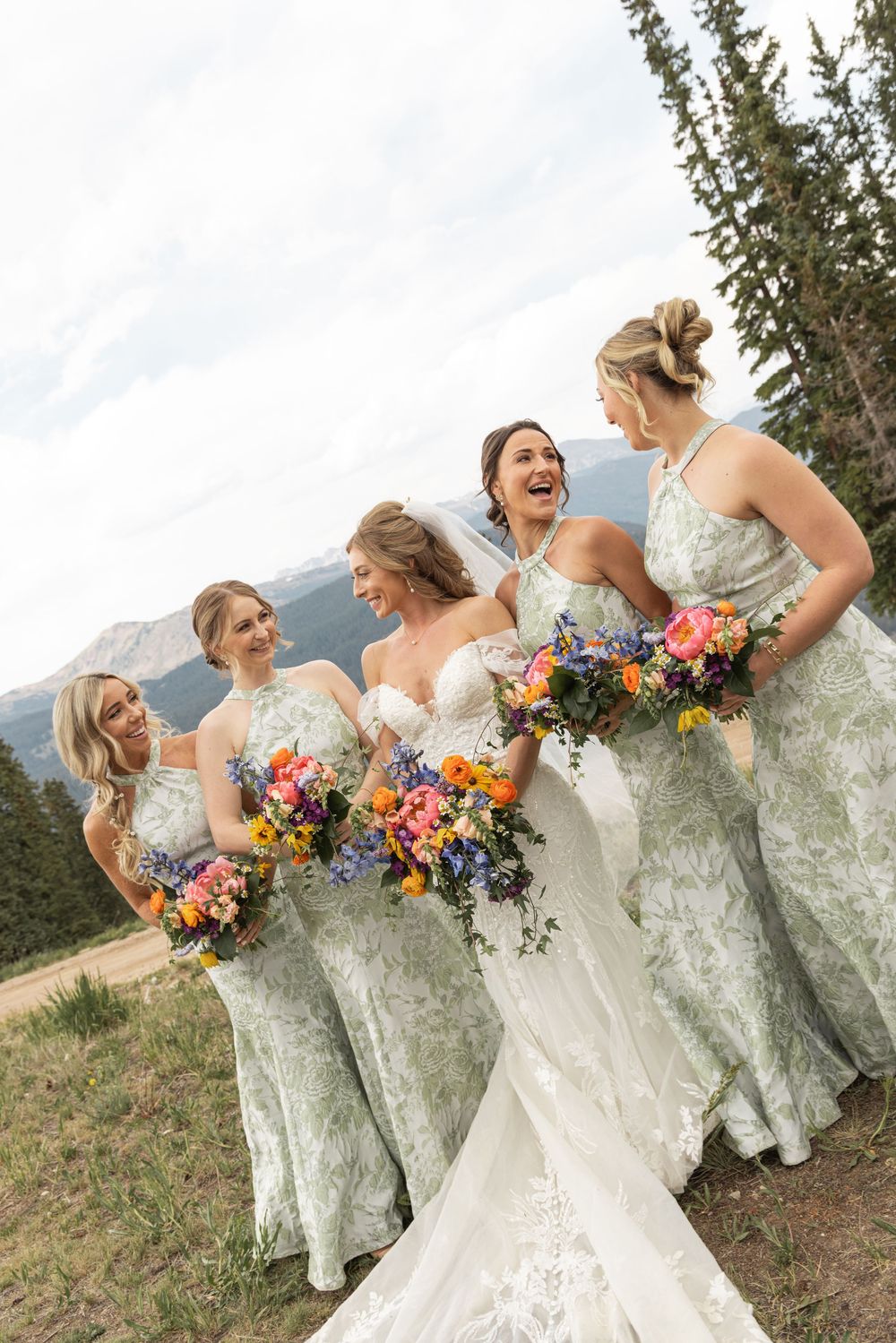 Bride and four bridesmaids in sage green dresses holding colorful bouquets laughing outdoors with mountain views.