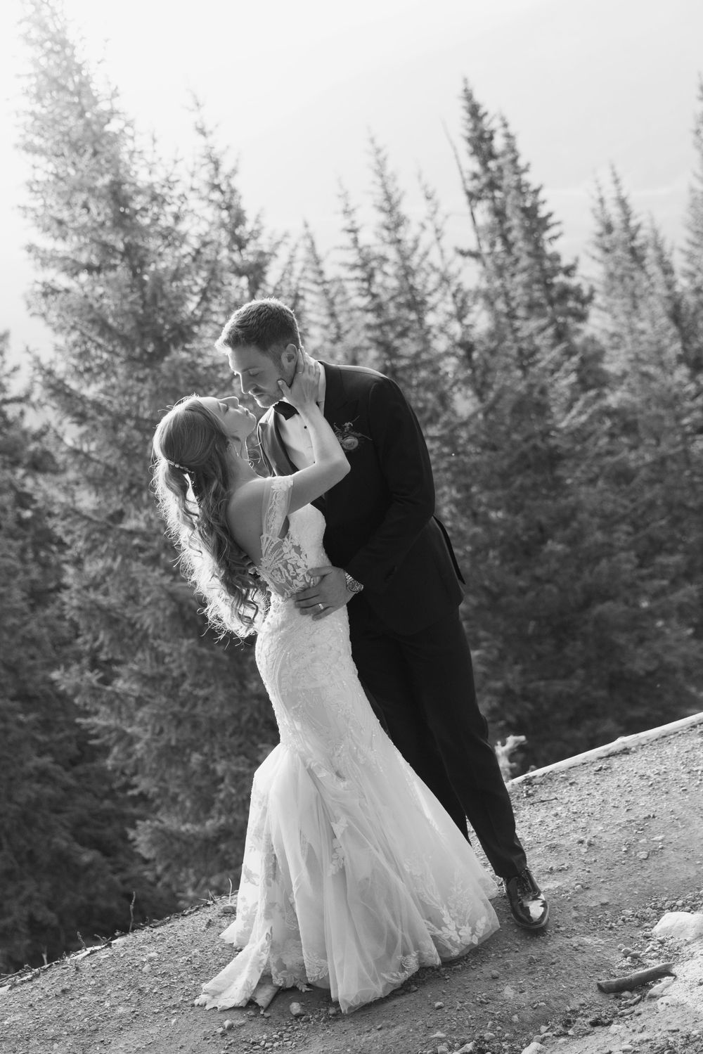 Black and white photo of a bride and groom sharing a romantic kiss outdoors surrounded by tall pine trees.