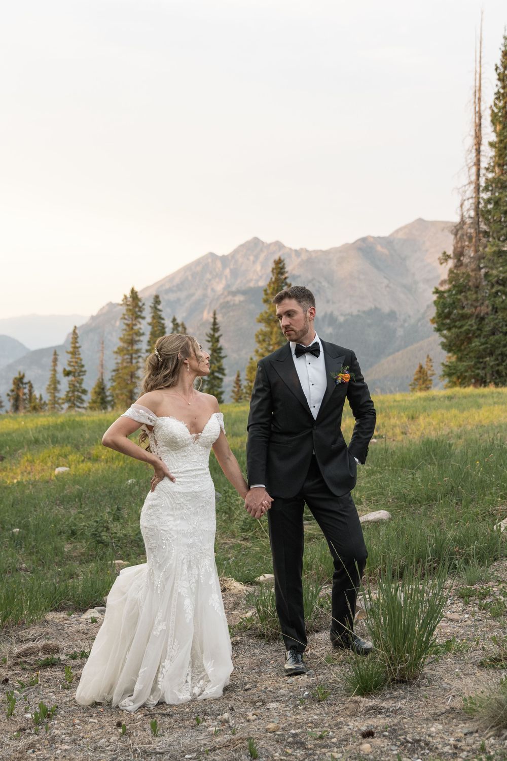 Bride in off-shoulder lace gown and groom in black tuxedo hold hands in a mountain meadow with pine trees.