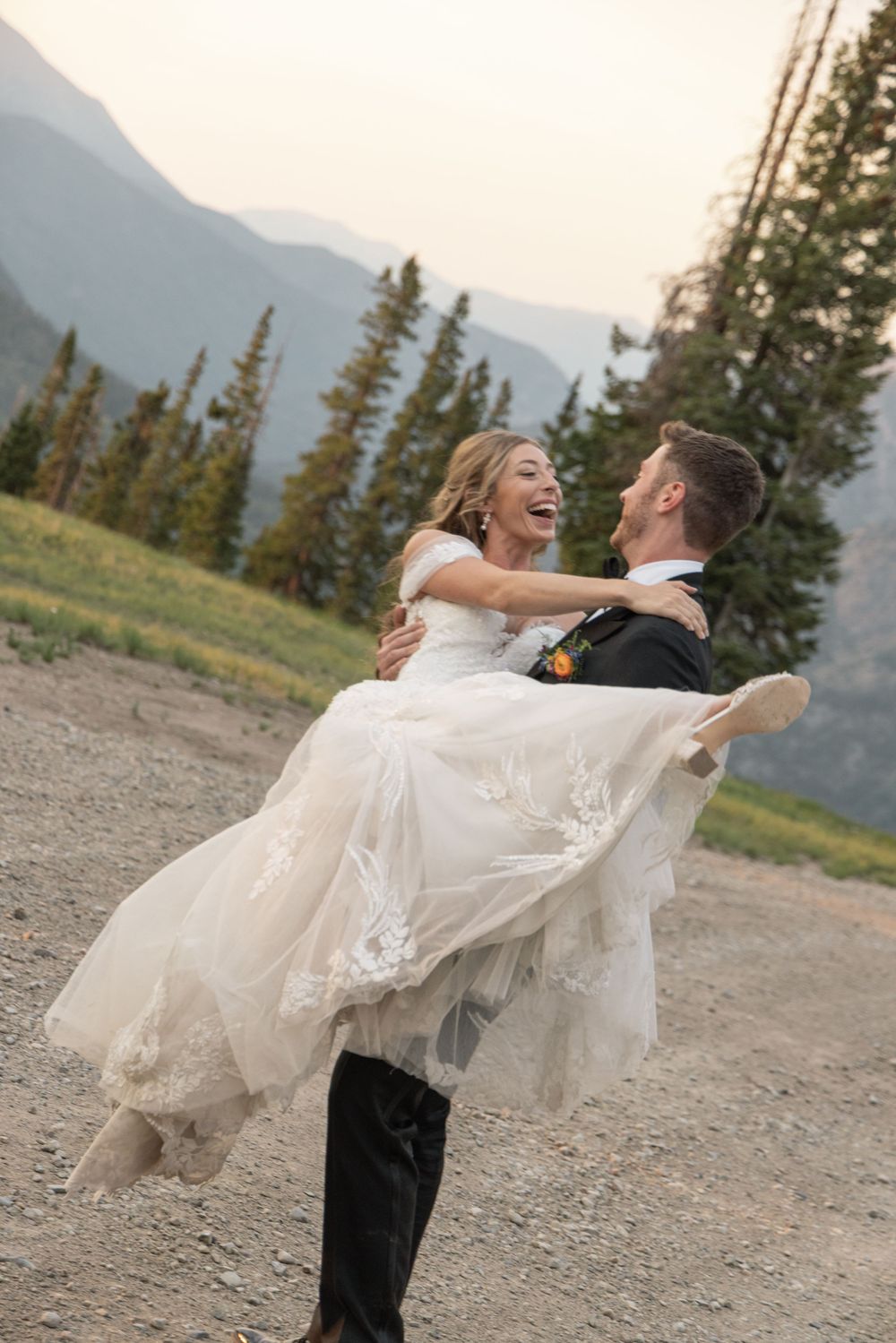 Joyful groom carrying laughing bride in white lace gown on mountain dirt path with evergreen trees backdrop.