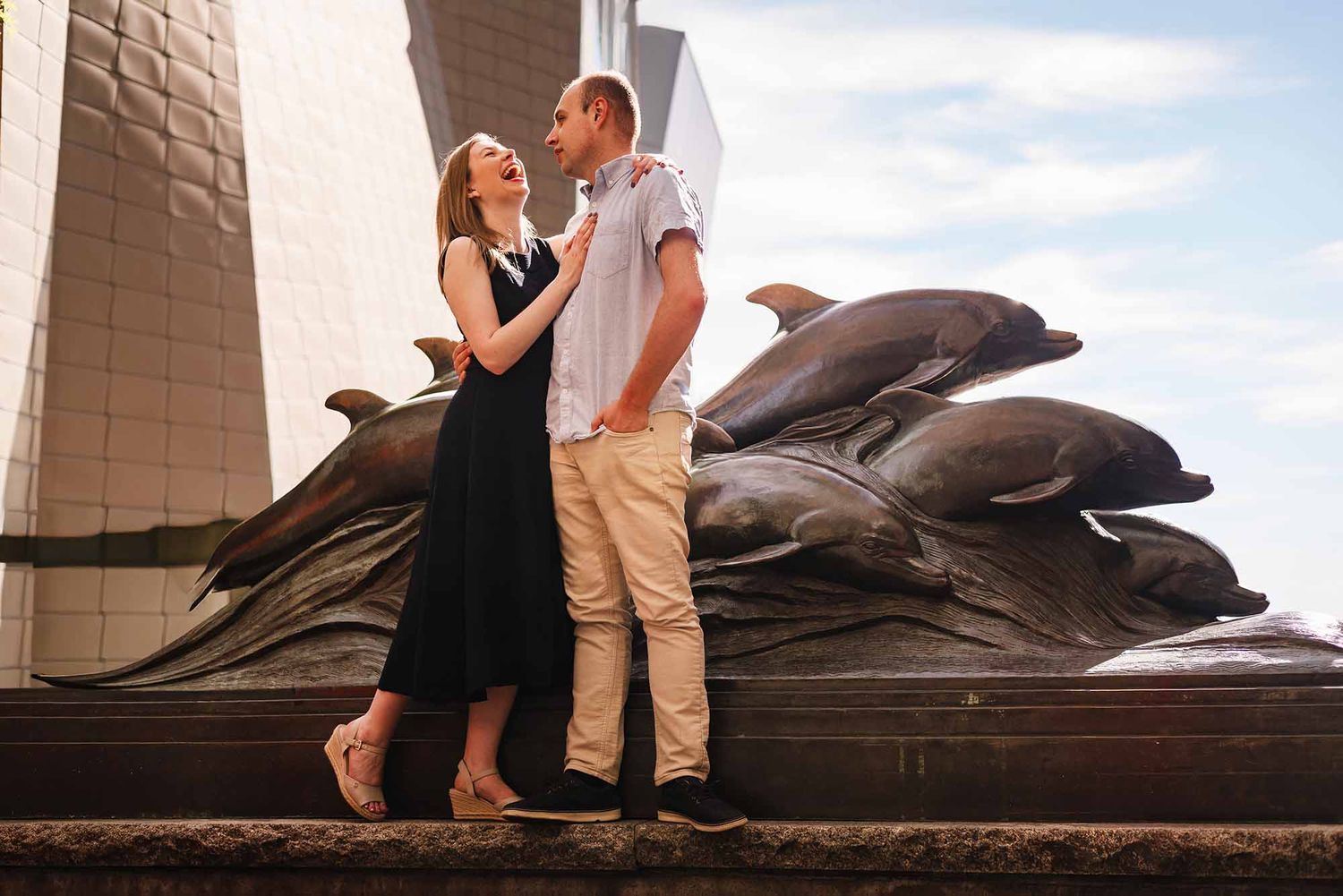 Engaged couple laughing together in front of the bronze dolphin statues at the New England Aquarium in Boston