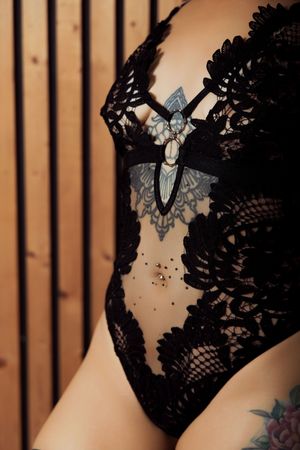 Black lace bodysuit with butterfly tattoo and floral details on a tattooed model against wooden backdrop.