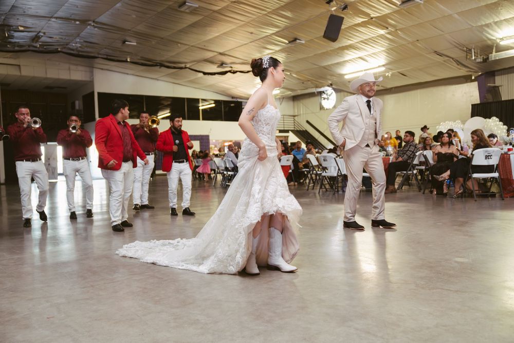 Bride in white gown walks with mariachi band in red jackets during wedding reception in large venue.