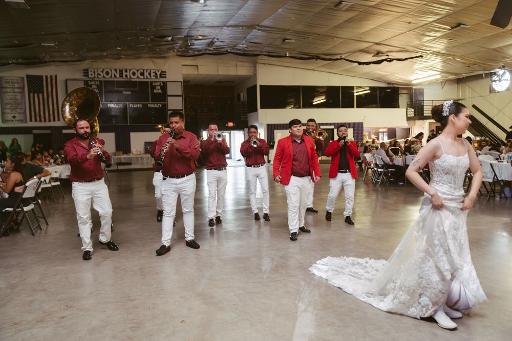 Bride in white lace gown leads groomsmen in red and maroon suits during wedding reception dance.