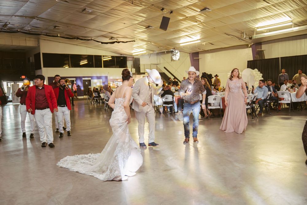 Bride in lace gown dances with groom in cowboy hat at wedding reception in large hall.