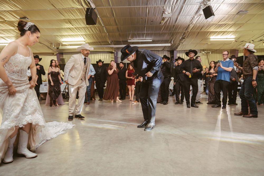 Groom in black cowboy hat bows to bride during lively wedding reception dance on concrete floor.