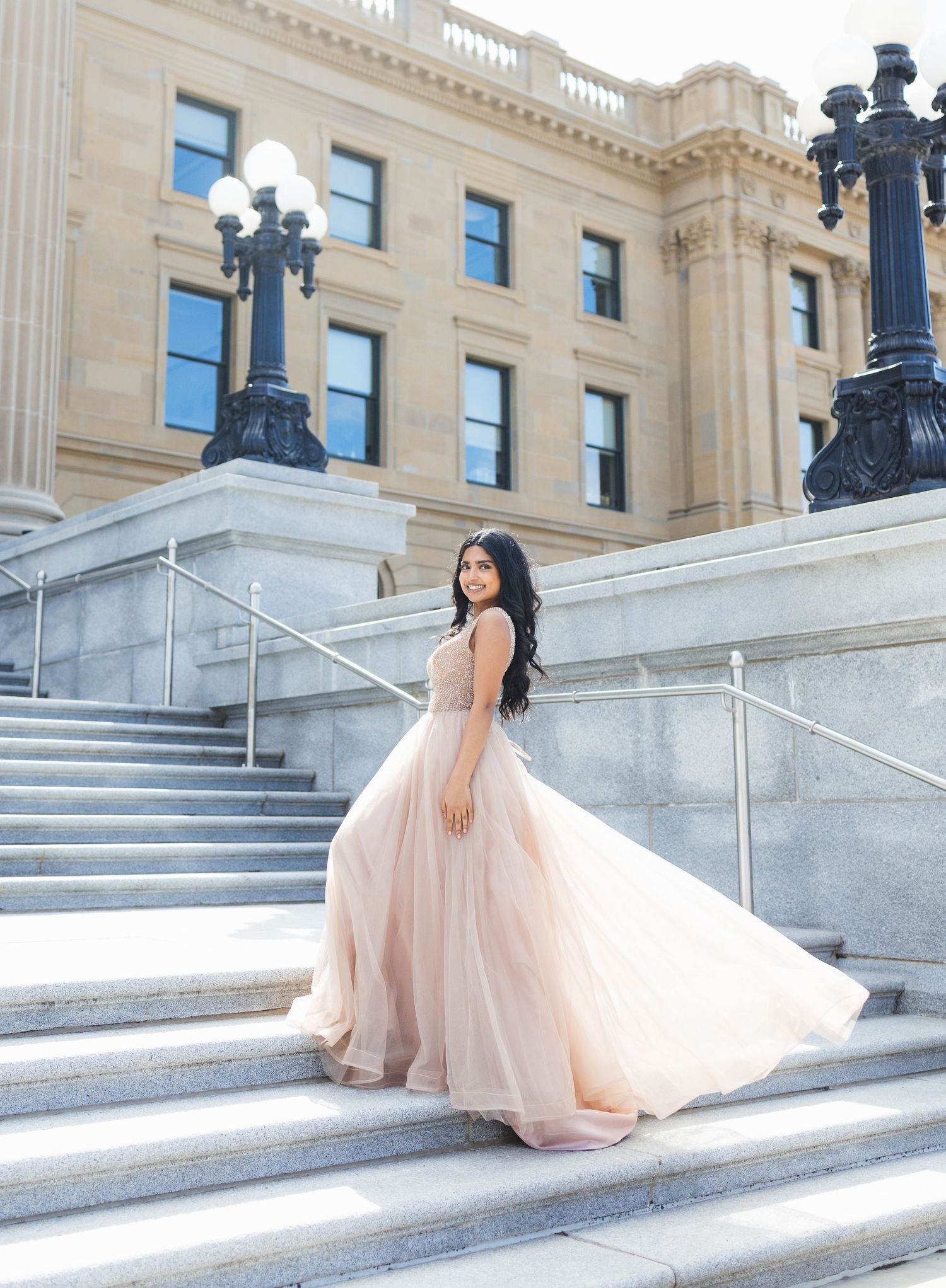 Young woman in flowing blush pink ball gown poses gracefully on stone steps of elegant neoclassical building.