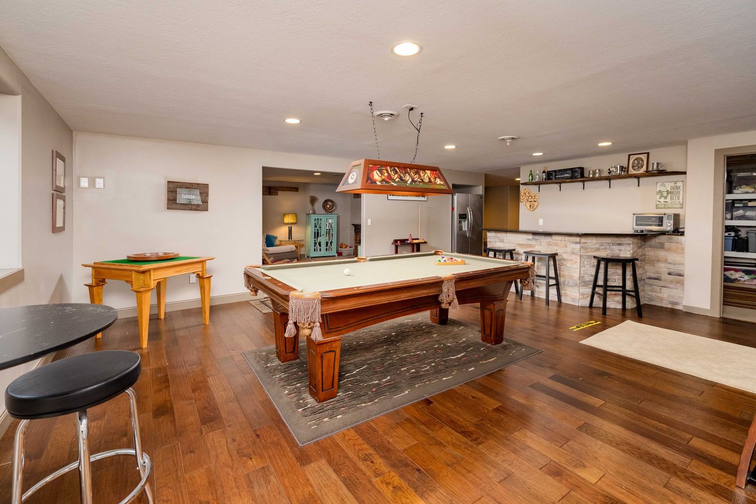 Spacious basement game room with a pool table, hardwood floors, bar area, and pendant lighting.