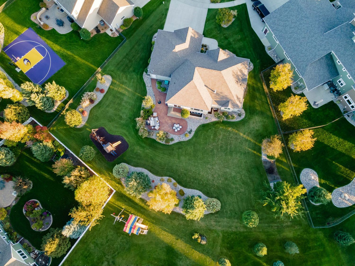 Aerial drone view of a large suburban home with lush green lawn, basketball court, and landscaped trees.