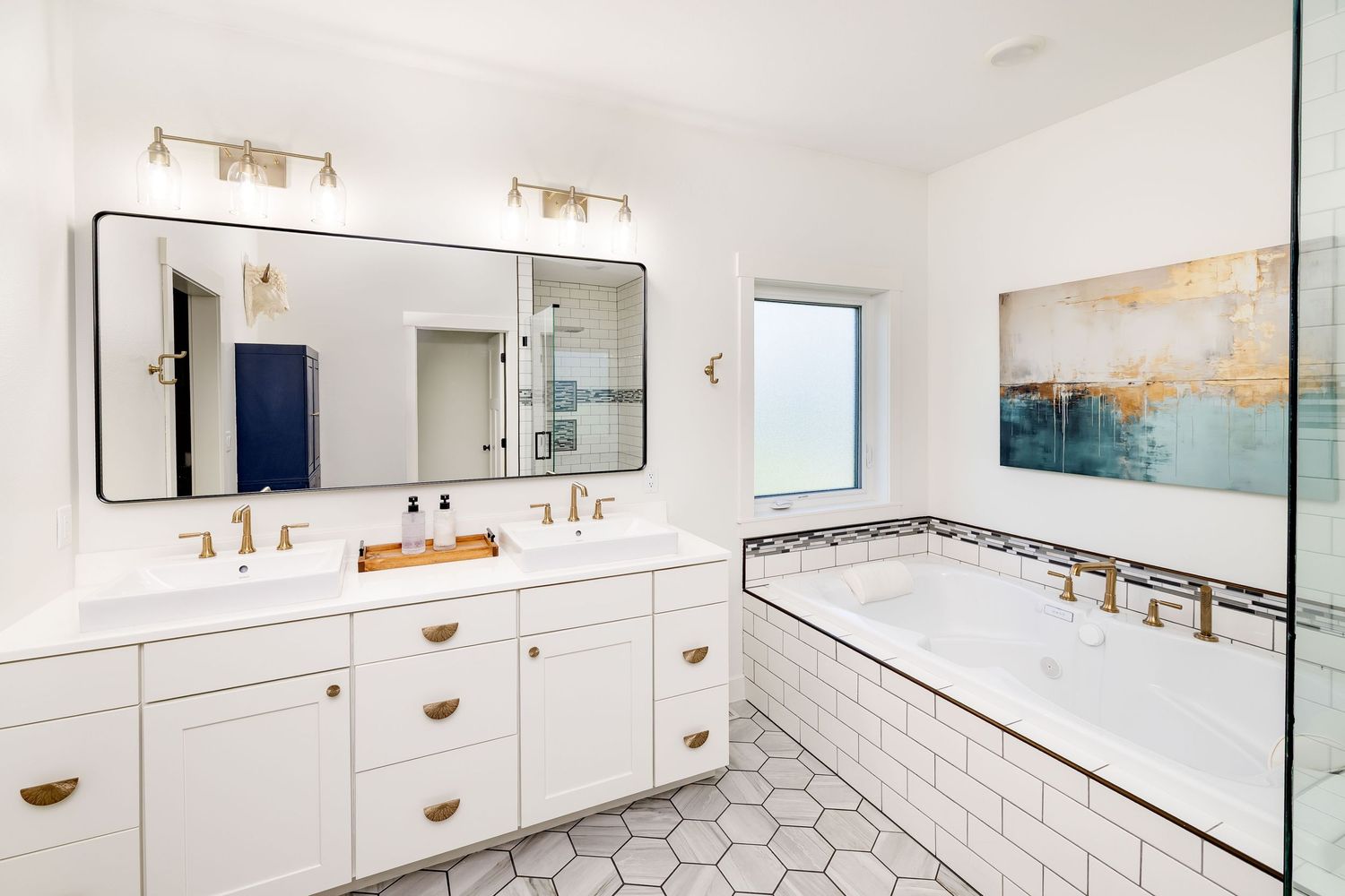 Modern white bathroom with double vanity, gold fixtures, hexagon tile floor, and tiled soaking tub.