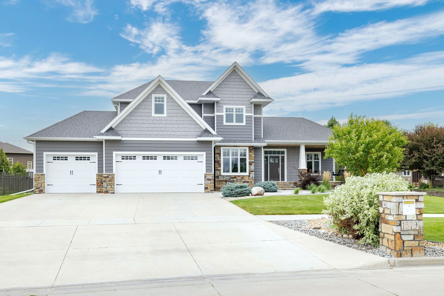 Craftsman-style home with gray siding, stone accents, three-car garage, and lush green landscaping.