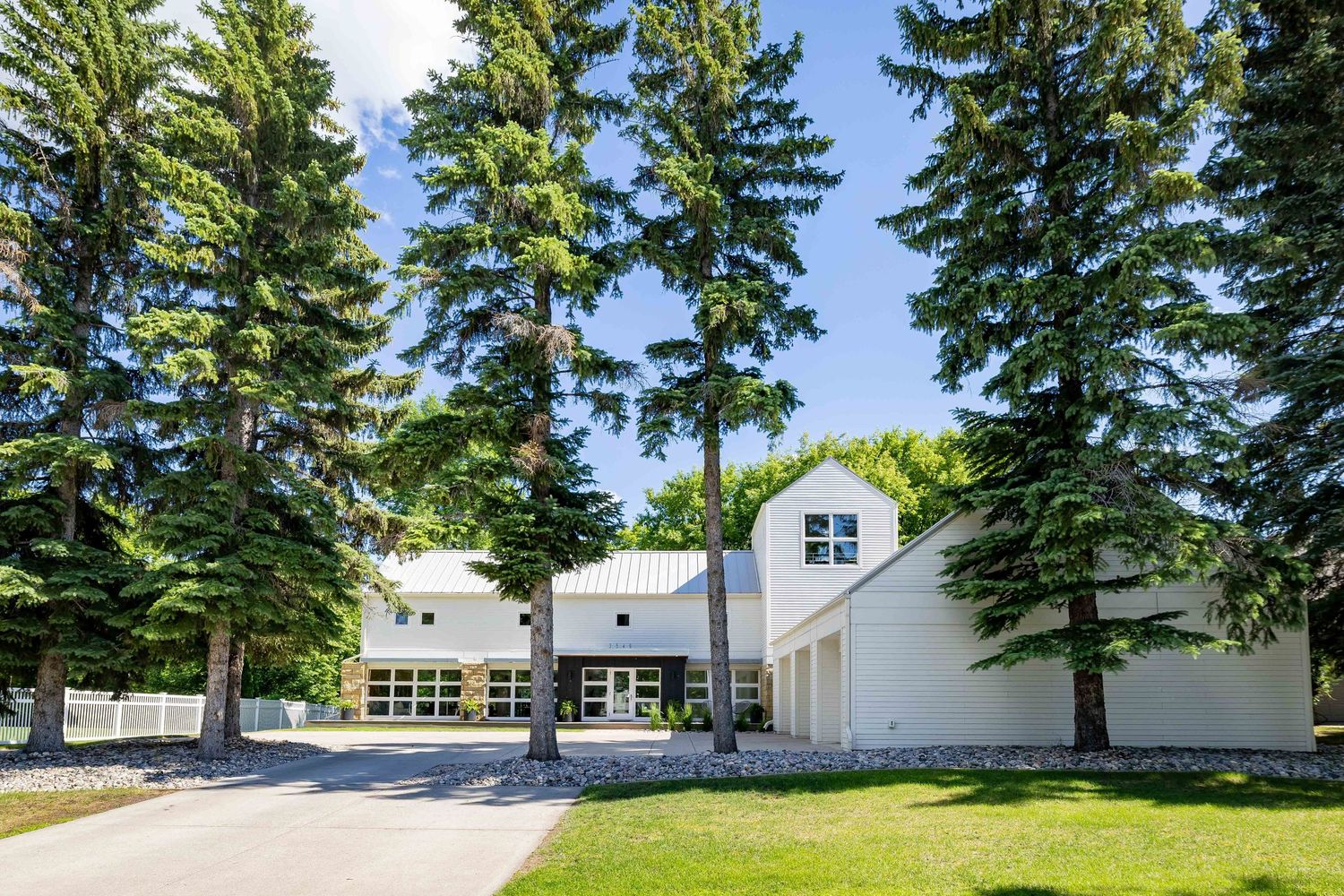 White two-story home surrounded by tall evergreen trees with a paved driveway and lush green lawn on a sunny day.