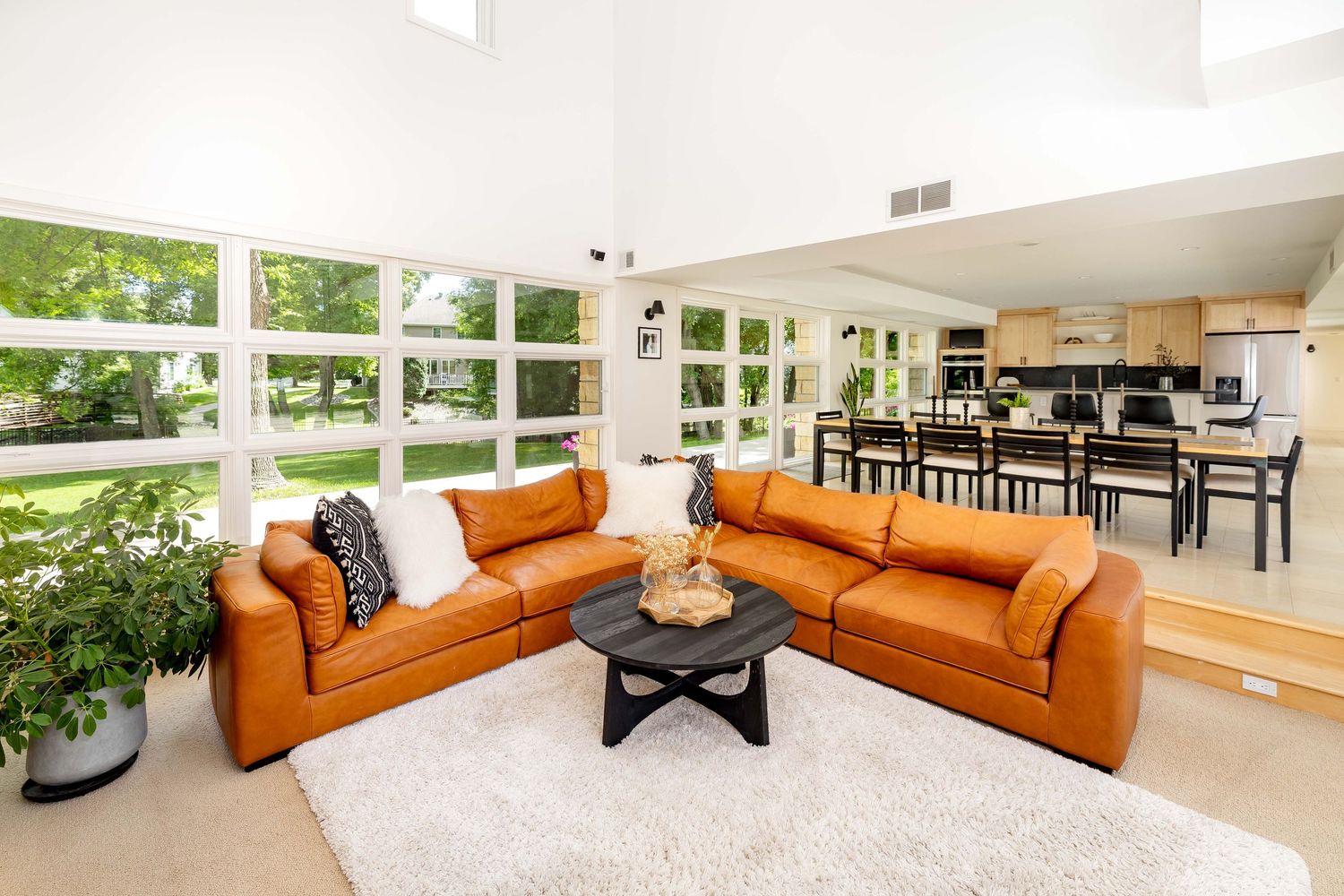 Bright open-concept living room with tan leather sectional, white shag rug, and views of lush greenery through large windows.