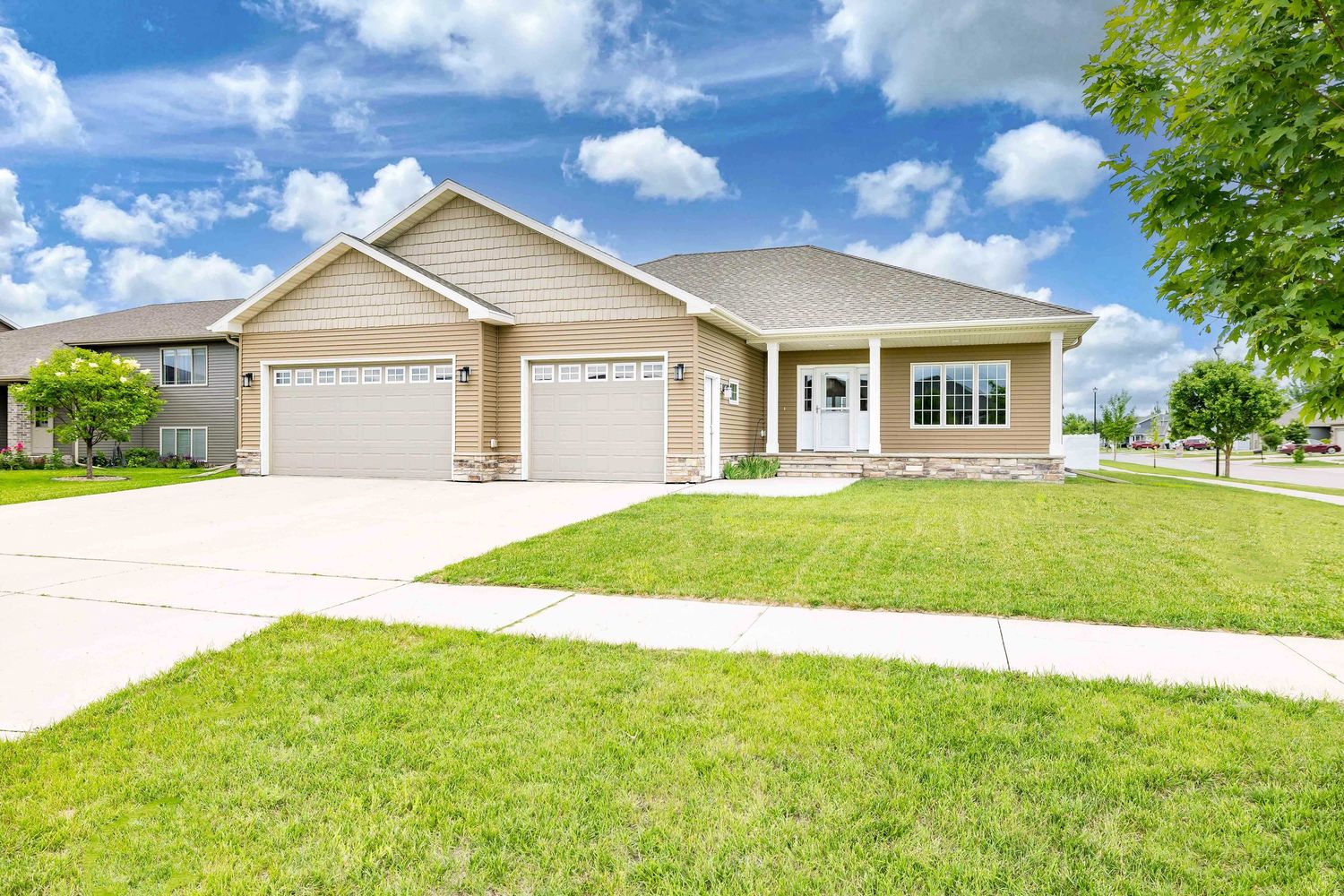 Ranch-style home with tan siding, two-car garage, covered porch, and lush green lawn under a bright blue sky.