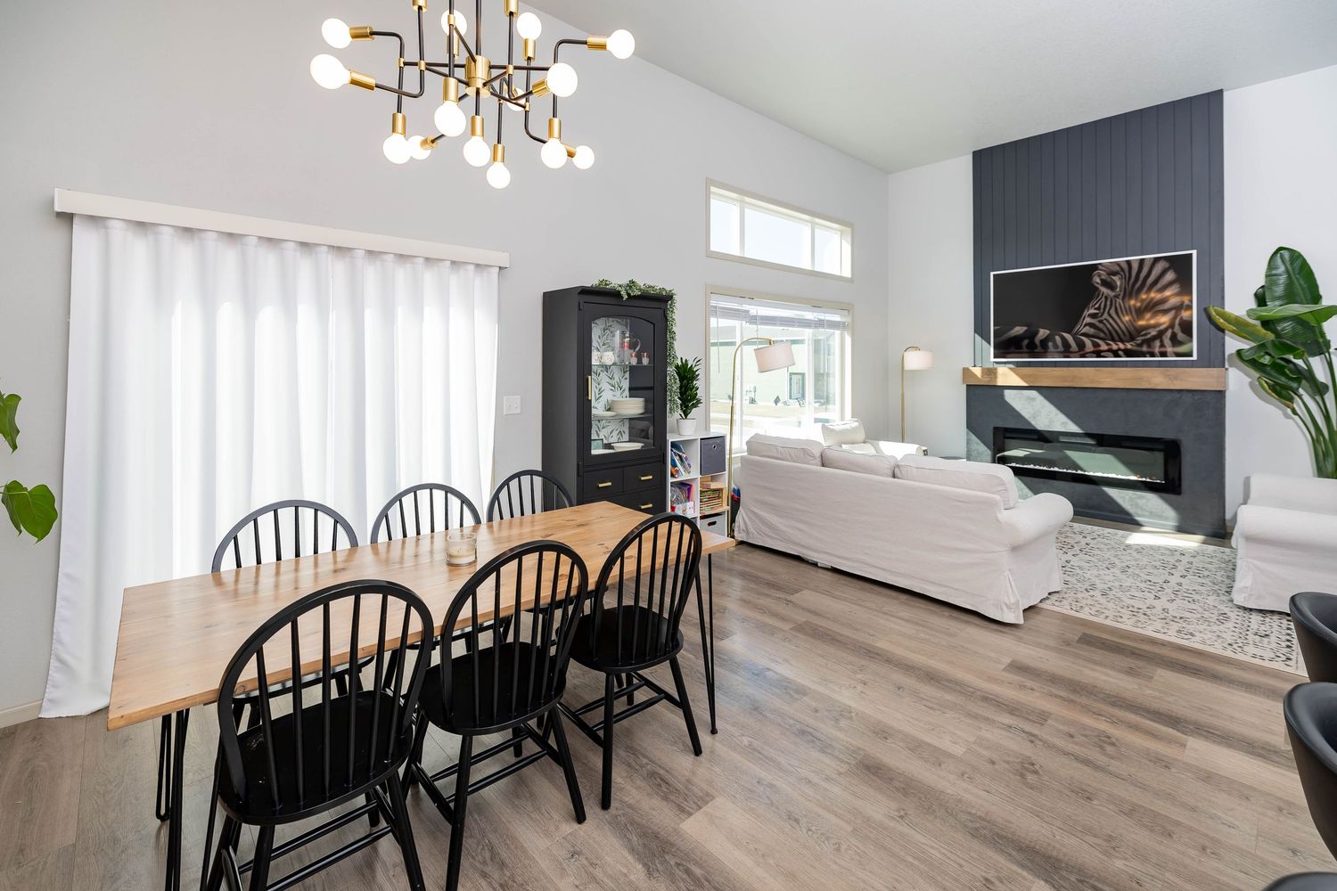 Modern open-plan dining and living room with wood floors, black chairs, white sofa, and dark shiplap fireplace wall.
