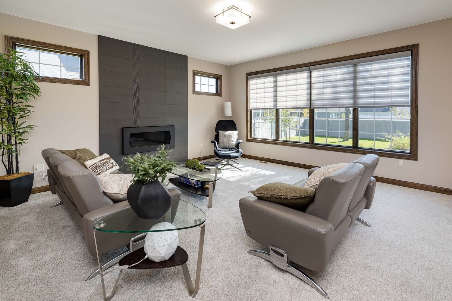 Modern living room with gray sofas, glass coffee table, tile fireplace, and large windows overlooking a green yard.