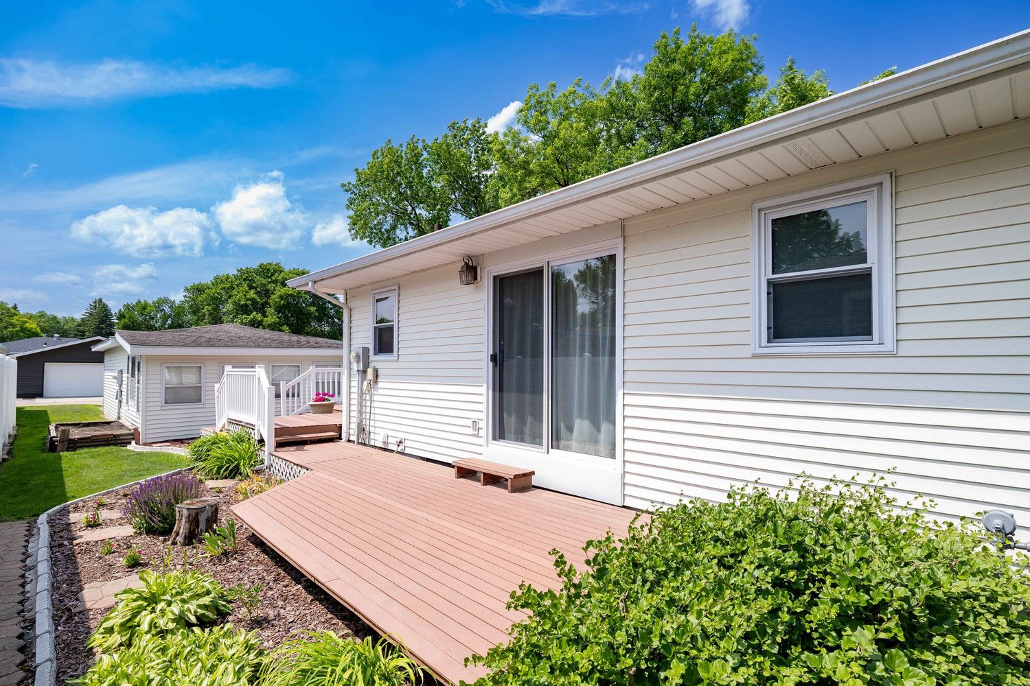 White ranch home exterior with wood deck, sliding glass door, lush green landscaping, and blue sky on a sunny day.