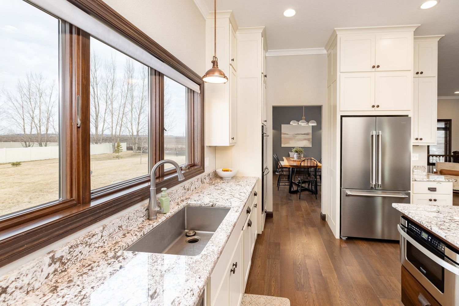 Modern kitchen with granite countertops, white cabinets, stainless steel fridge, and large windows overlooking a scenic landscape.