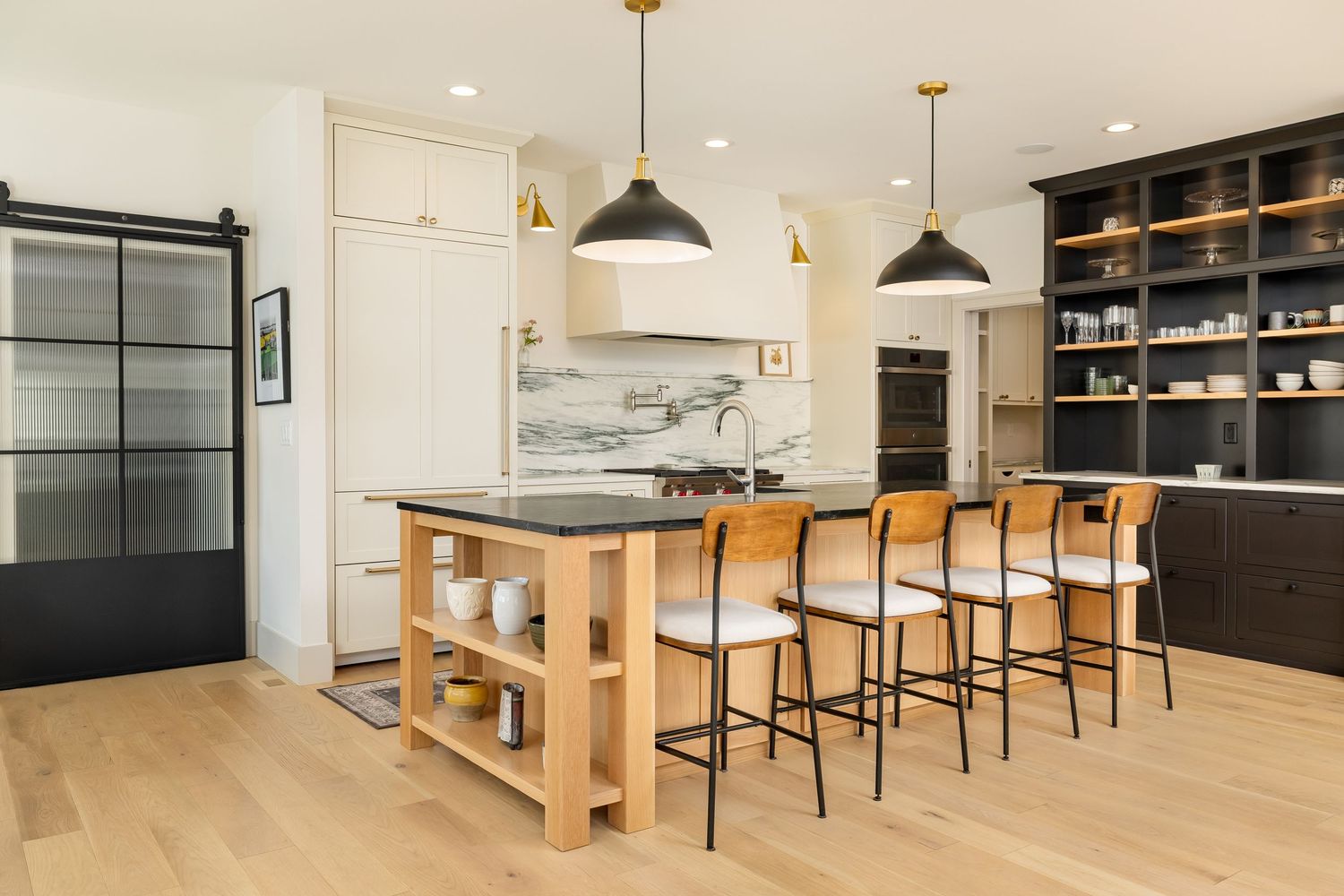Modern kitchen with black pendant lights, wood island, white bar stools, dark shelving, and light hardwood floors.