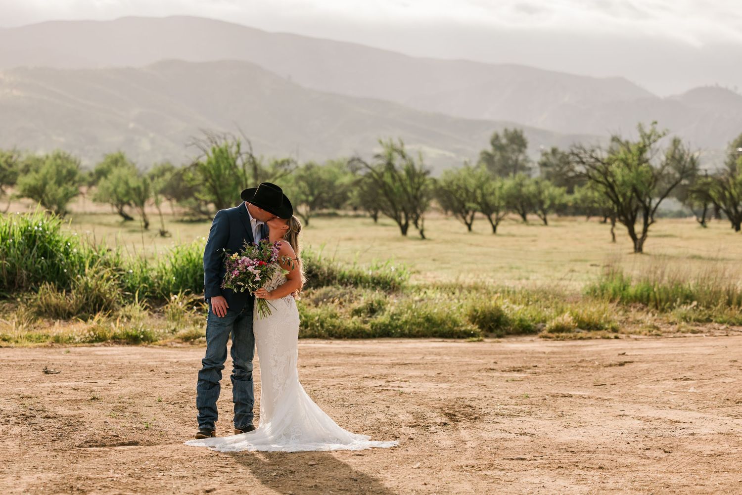 Bride and groom kissing in rustic field with mountain backdrop, groom in cowboy hat and jeans, bride in white gown.