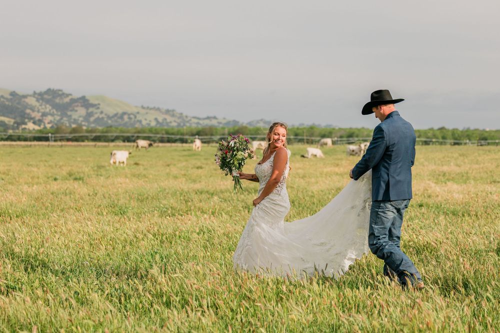 Bride and groom walking through green field, man in blue suit and cowboy hat holding wedding dress train.