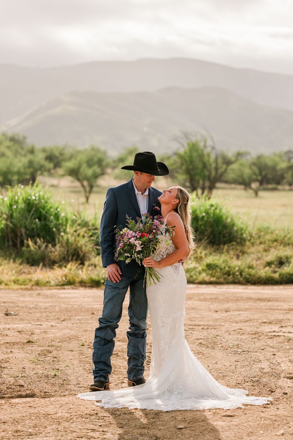 Bride and groom share a romantic moment outdoors, with mountains and greenery behind them, as she holds a colorful bouquet.