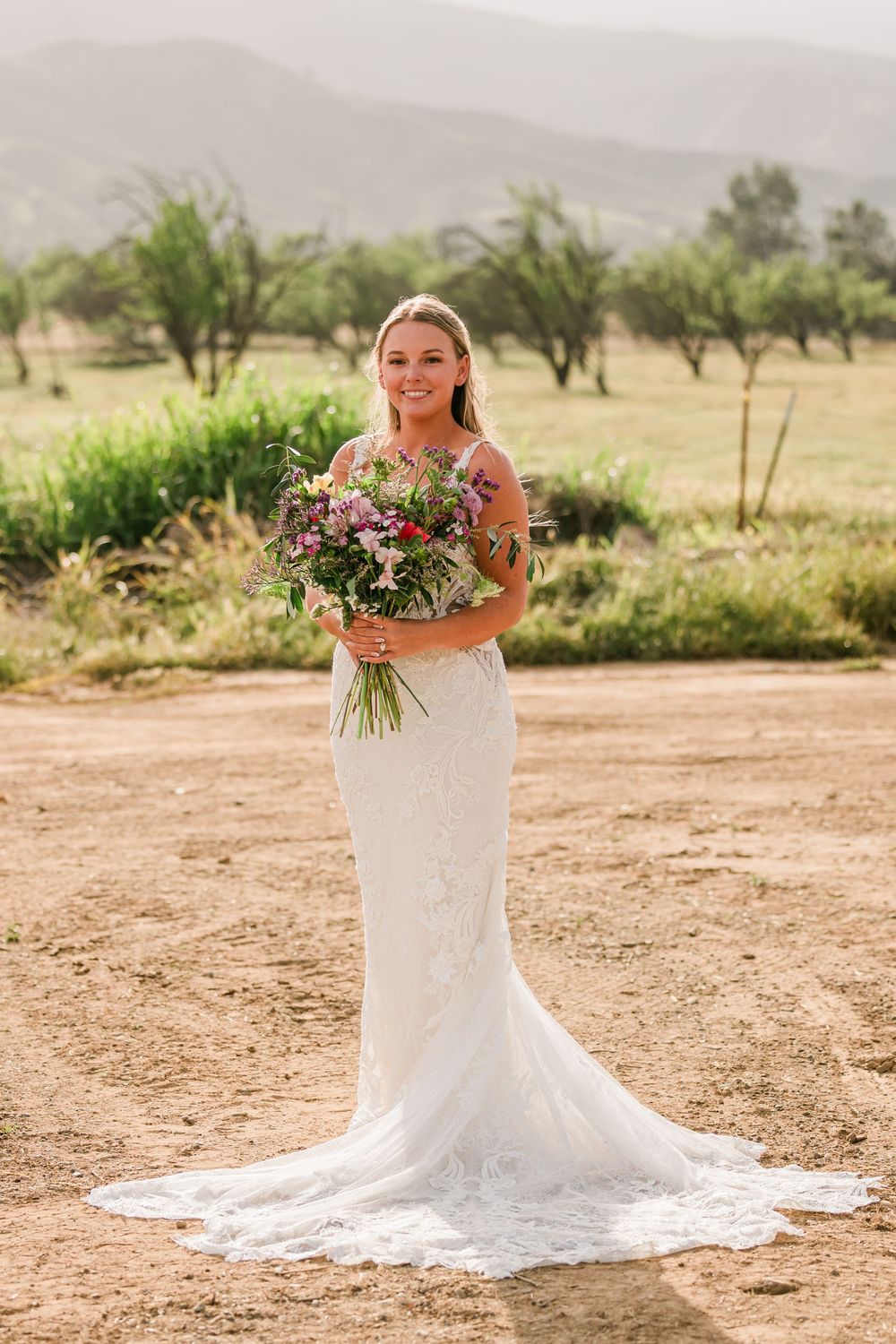 Bride in white lace mermaid gown holding colorful bouquet outdoors with mountains in background.