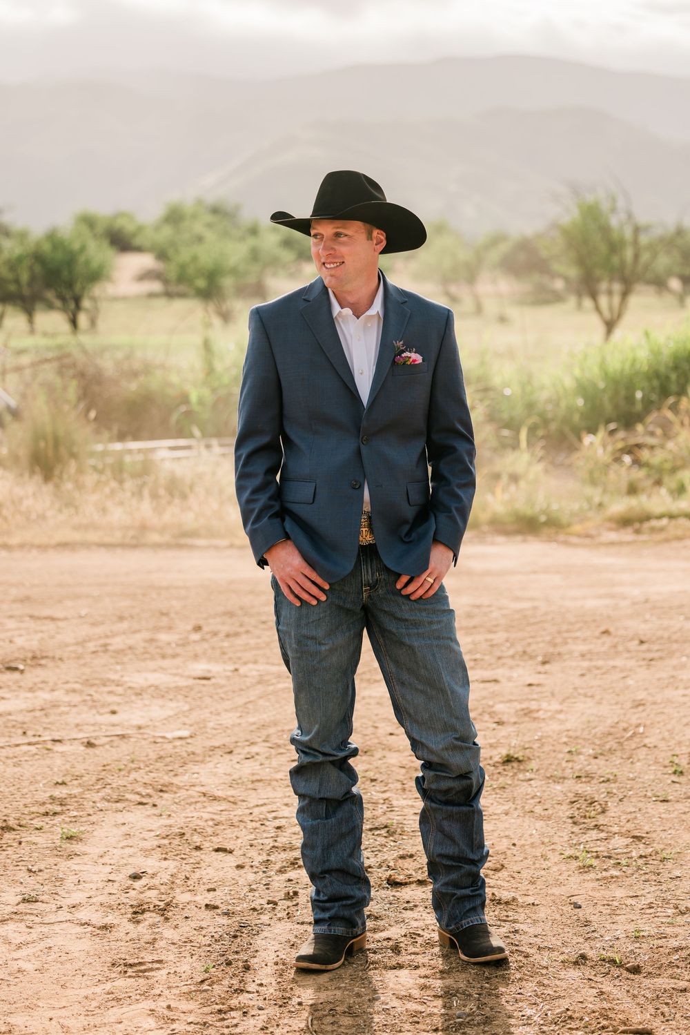 Groom in blue blazer, jeans, cowboy hat and boots standing on dirt path with mountains behind.