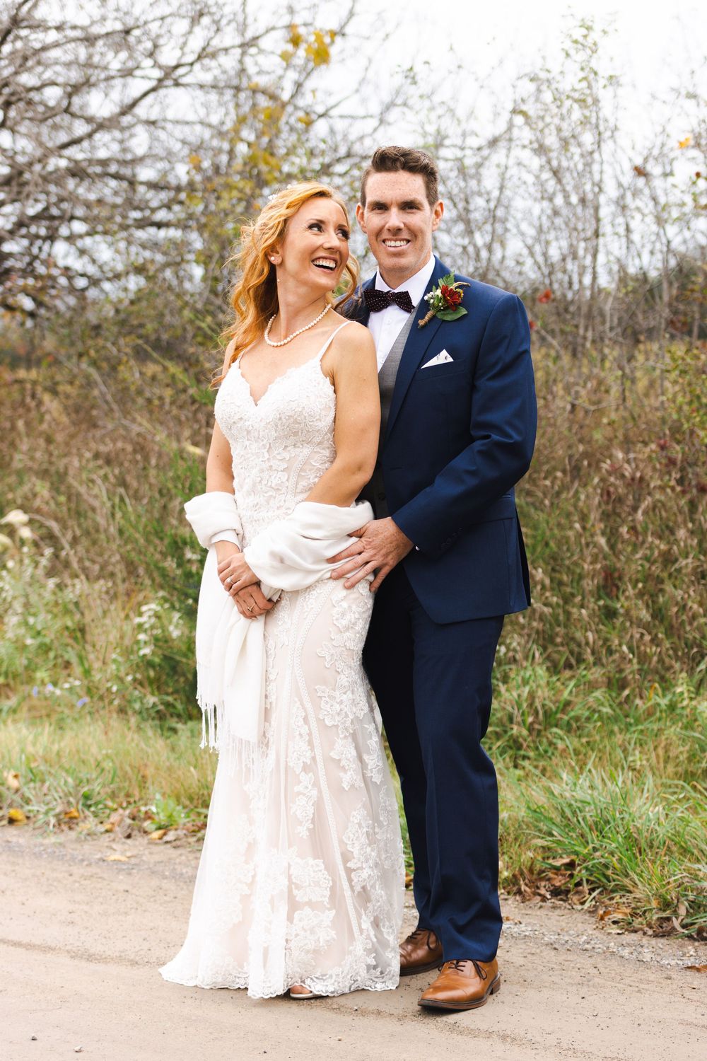 Smiling bride in lace gown and groom in navy tuxedo posing outdoors on a fall wedding day.