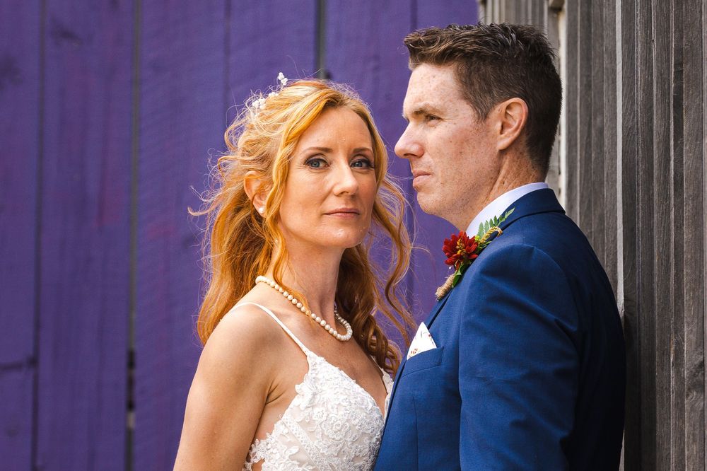 Bride and groom posing against a purple wooden wall, wearing wedding attire with a red boutonniere.