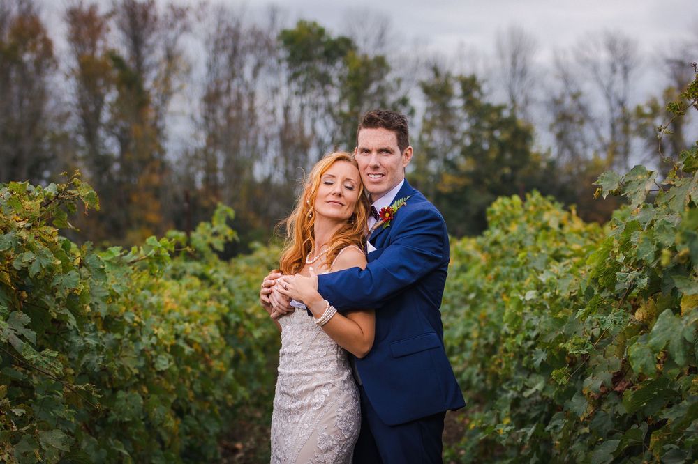 Bride and groom embracing in a lush vineyard, wearing wedding attire, surrounded by green grapevines in autumn.