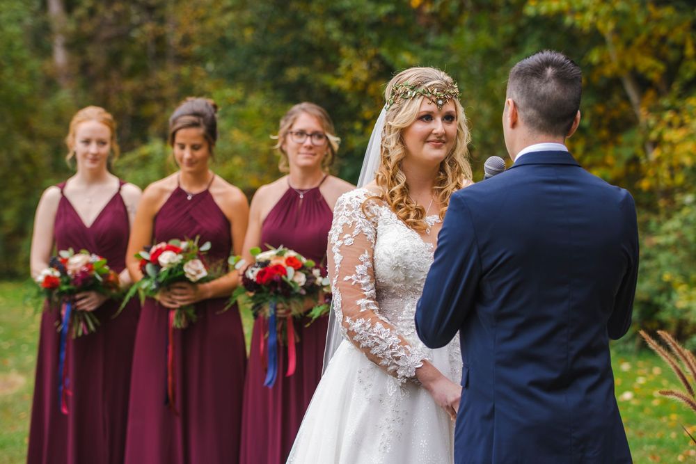 Bride in lace gown with floral crown exchanges vows with groom as bridesmaids in burgundy dresses hold bouquets outdoors.