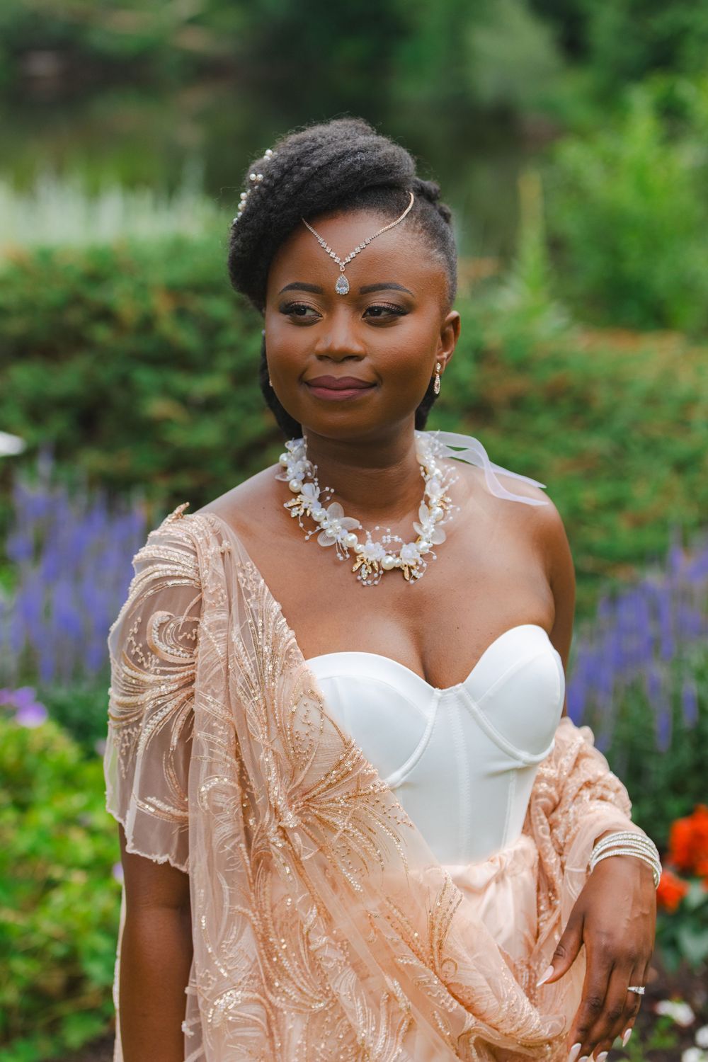 Elegant Black bride in white corset gown with beaded necklace and shawl posing in lush garden setting.