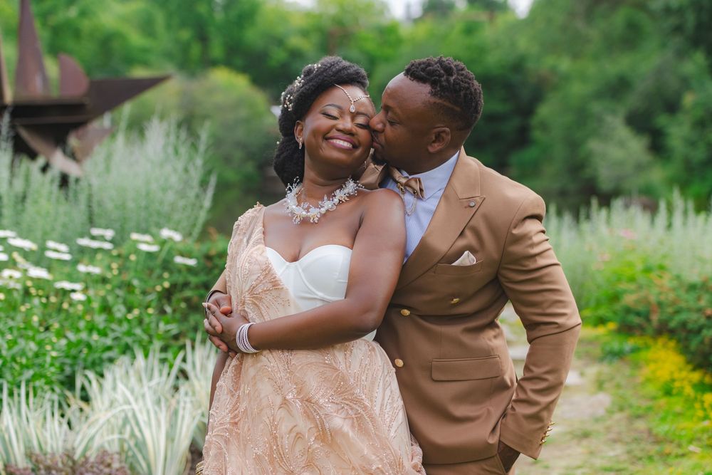 A joyful groom in a tan suit kisses his bride in a blush gown outdoors at a garden wedding.