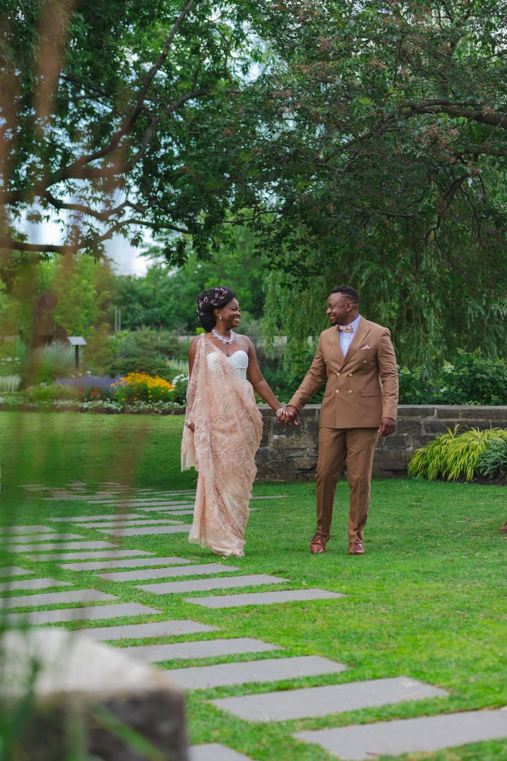 Couple in elegant wedding attire holding hands while walking along a stone path in a lush green garden.