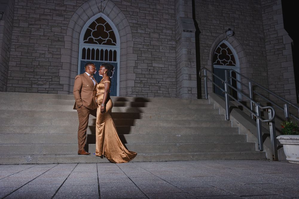 Elegant couple in formal attire posing at night outside a stone church with arched windows and steps.