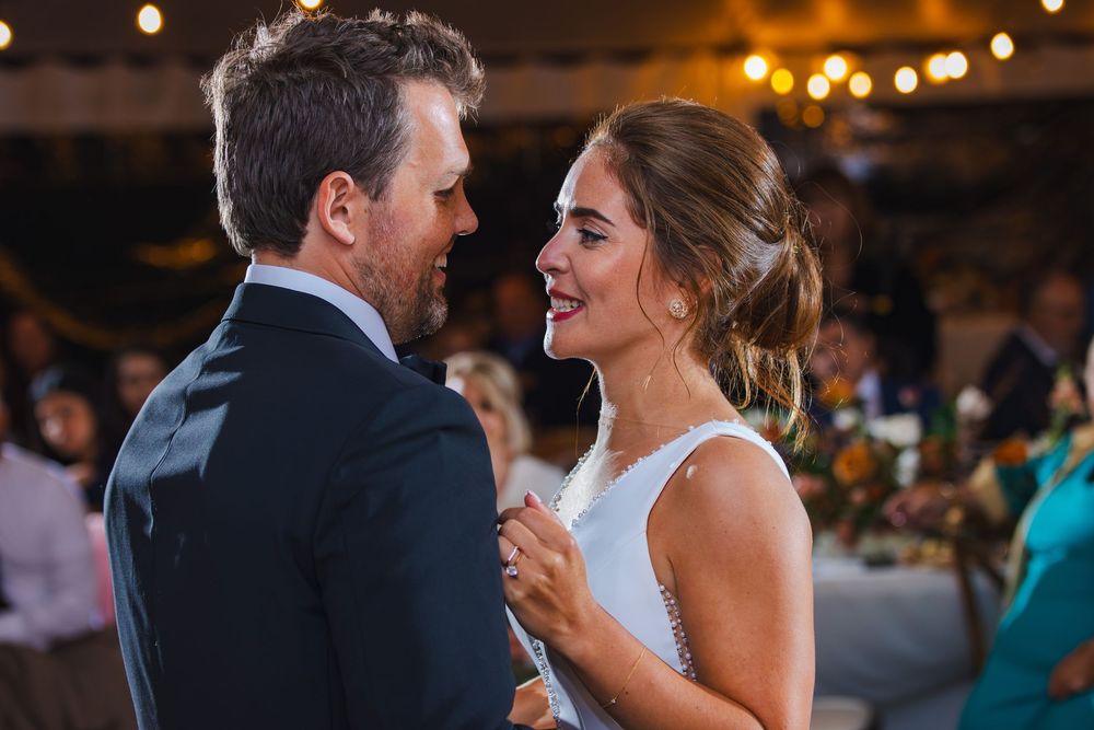 Bride and groom share their first dance, smiling joyfully under warm string lights at a wedding reception.