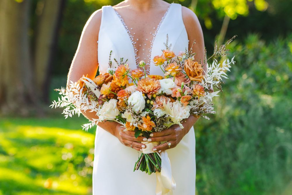Bride in white gown holding a lush bouquet of orange, white, and peach flowers with greenery outdoors.