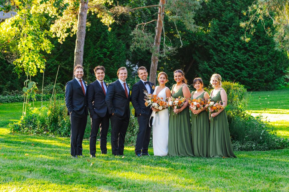 Wedding party posing outdoors with groomsmen in suits and bridesmaids in sage green gowns holding bouquets.