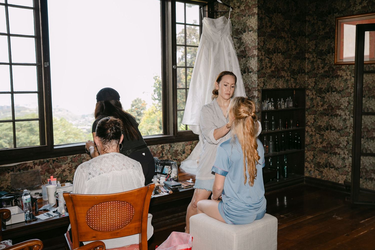Bride in white robe gets hair styled by hairdresser in hotel room before wedding ceremony.