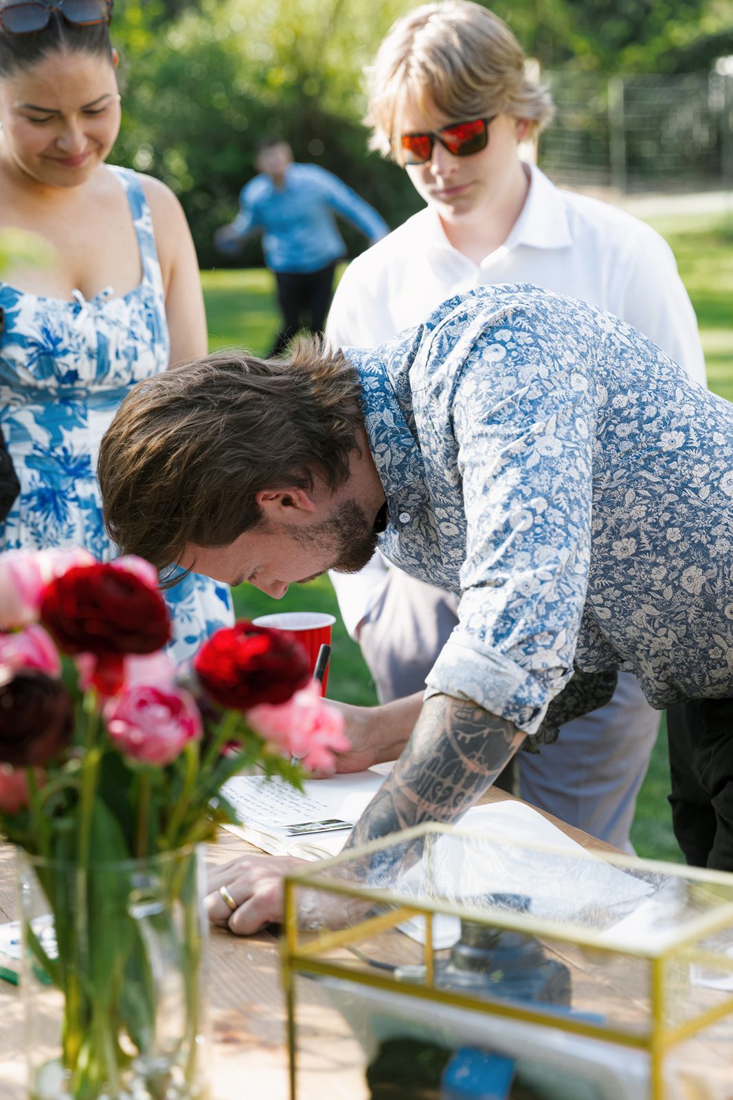 Outdoor garden party with guests mingling under white patio umbrellas surrounded by lush green trees on a sunny day.