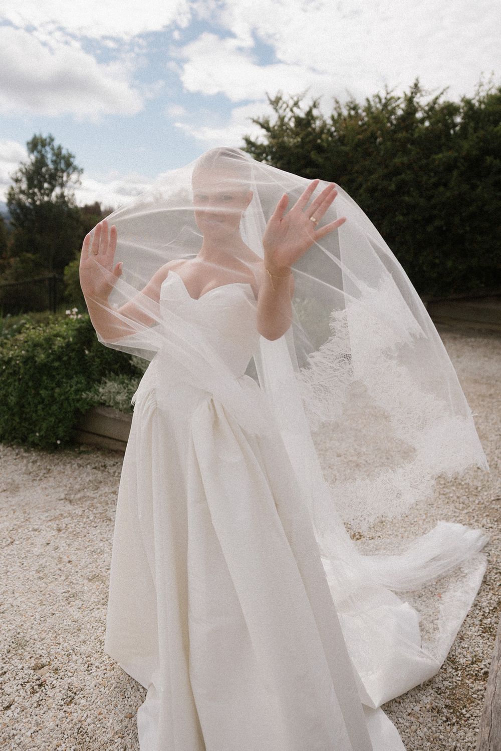 Bride in strapless white ball gown with flowing veil blowing over her face outdoors.