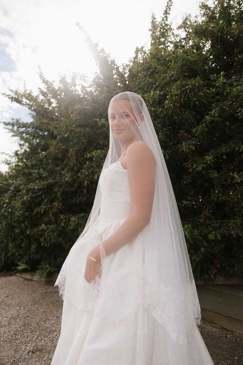 Bride in white gown and lace veil poses outdoors with sunlight filtering through green trees behind her.