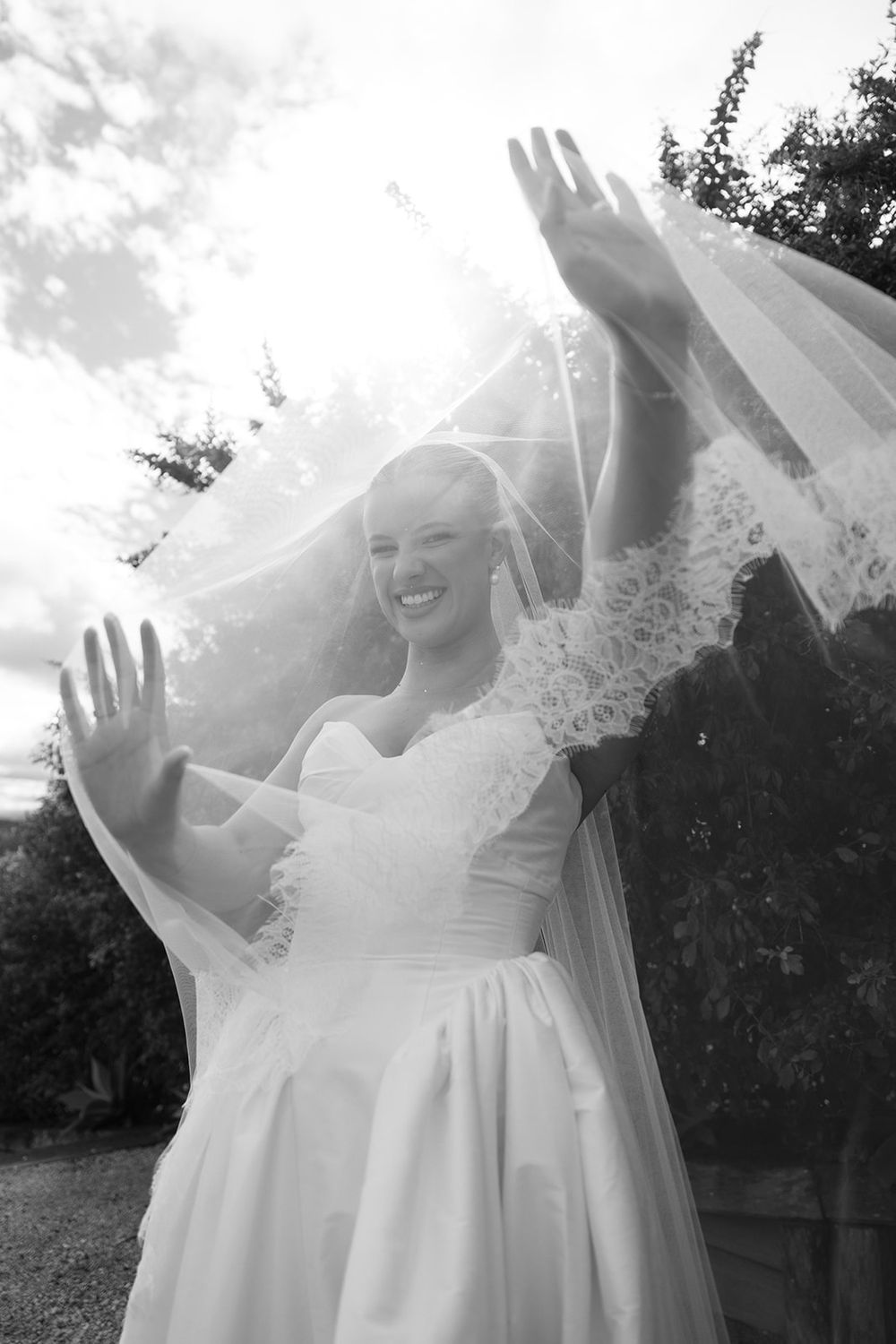 Black and white photo of a smiling bride in white gown and veil, arms raised joyfully outdoors on her wedding day.