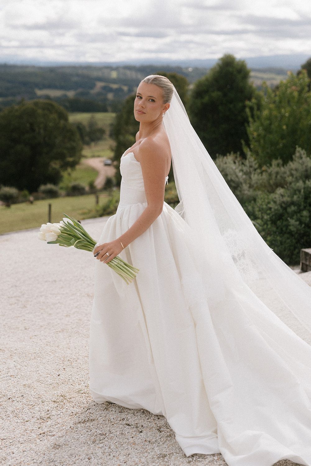 Bride in strapless white ball gown with flowing veil holds white tulip bouquet on gravel path with scenic countryside backdrop.